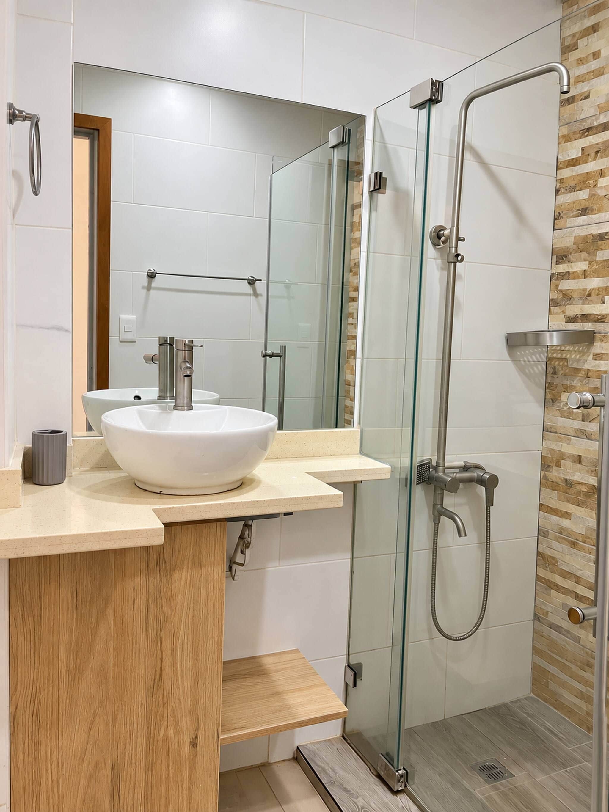 Een moderne badkamer met een glazen douchecabine, een witte wastafel en een houten opbergmeubel.