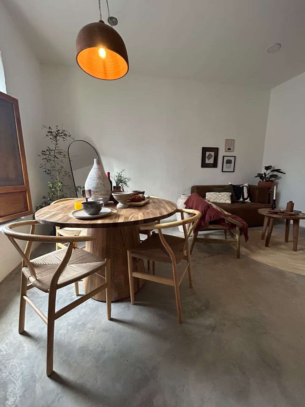 A round wooden dining table with chairs, a hanging pendant light, a floor lamp, a sofa, a small side table, and a potted plant.