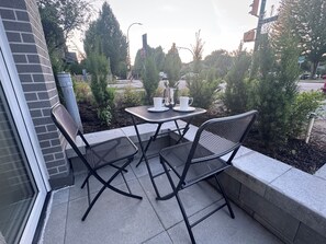 Un patio al aire libre con dos sillas, una mesa, un muro de ladrillo y vista a la calle con semáforos y árboles.