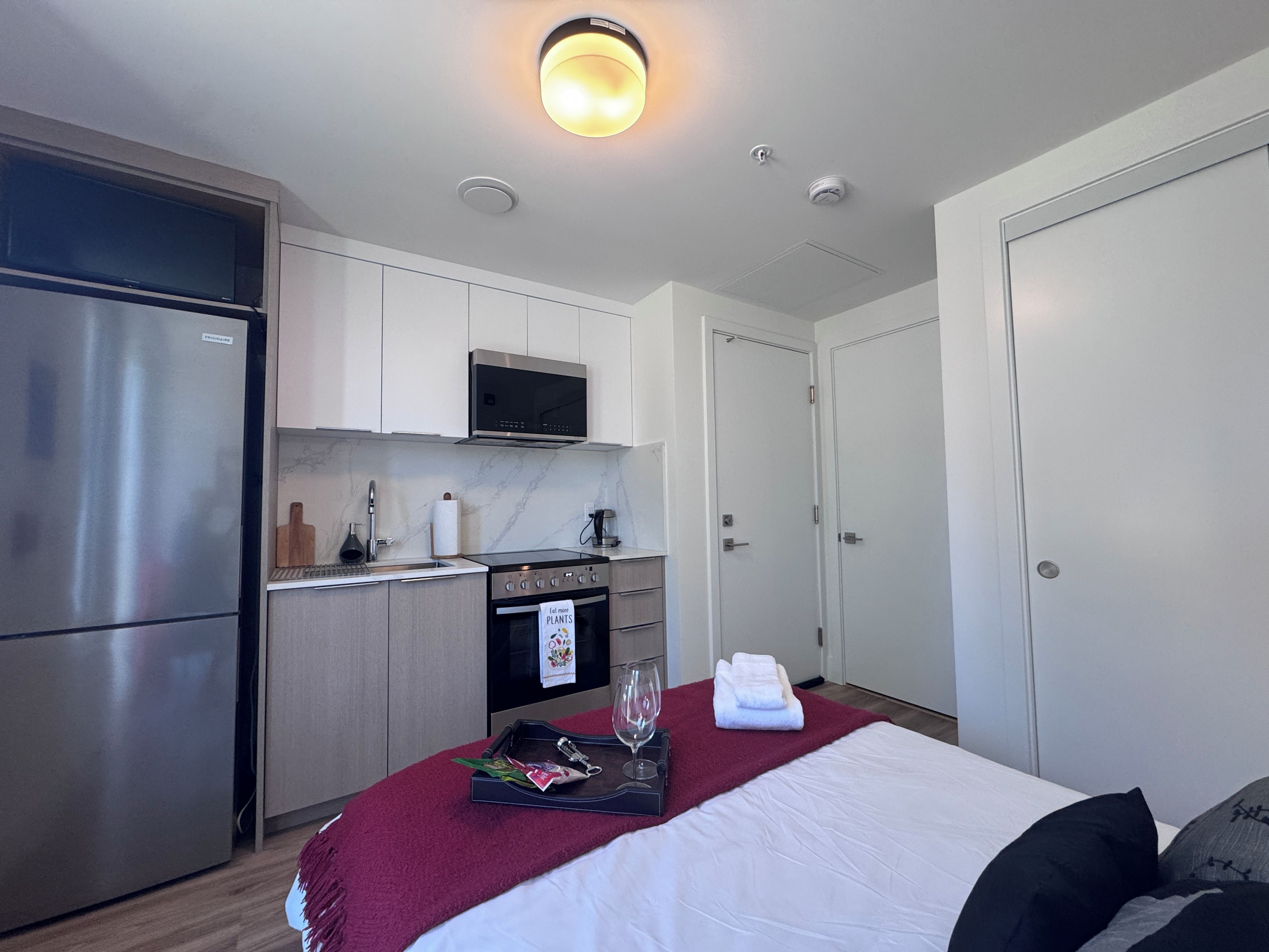 A modern hotel room with a kitchenette, a bed with a red blanket, and a glass of water on a tray.