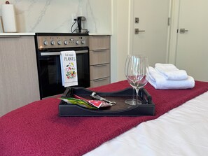 Habitación de hotel con una cama, un servicio de mesa con una copa de vino y una botella, una toalla y una zona de cocina con cafetera.
