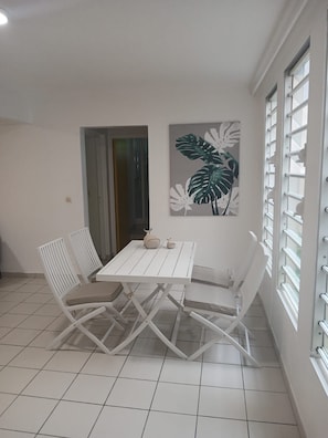 Un comedor con una mesa y sillas blancas, un cuadro de hojas tropicales y una ventana con persianas.