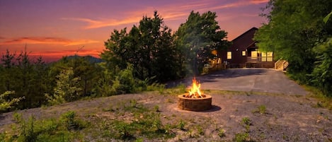 A house with a fire pit in the foreground and a driveway leading to the entrance.