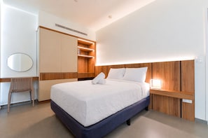 Una habitación de hotel moderna con una cama grande, cabecera de madera, mesita de noche con lámpara, armario empotrado y un espejo redondo.
