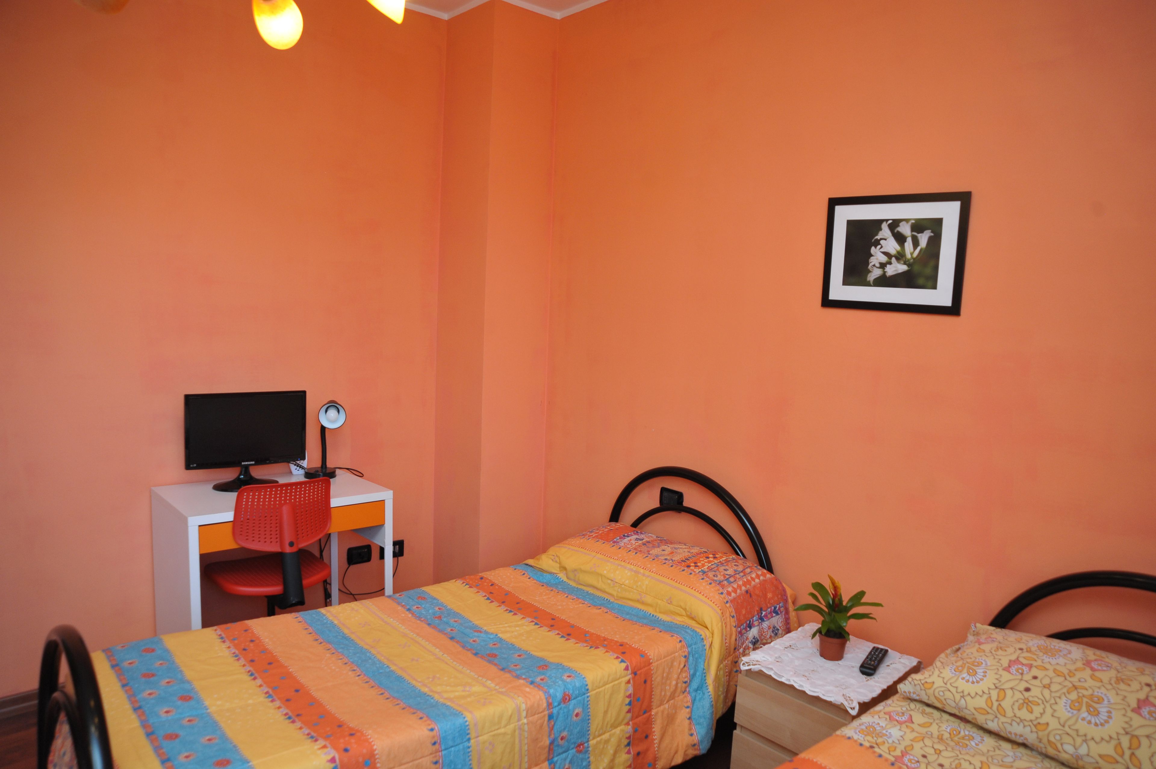 Una camera da letto con due letti singoli, una scrivania con un computer, una sedia, un quadro appeso alla parete e una piccola pianta in vaso.