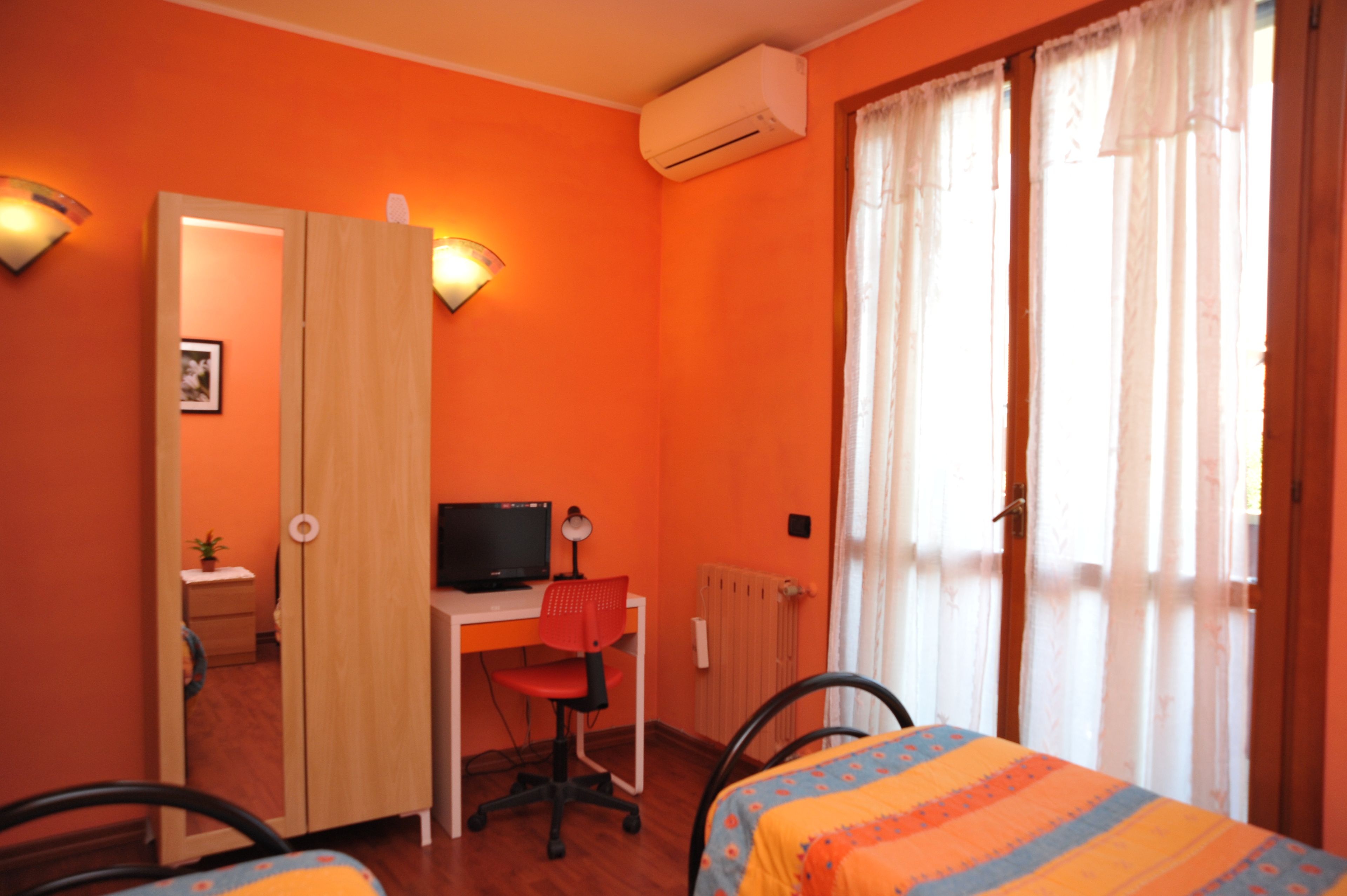 Una stanza con pareti arancioni, una scrivania con un computer, due sedie, un letto con coperta a righe e una finestra con tende.