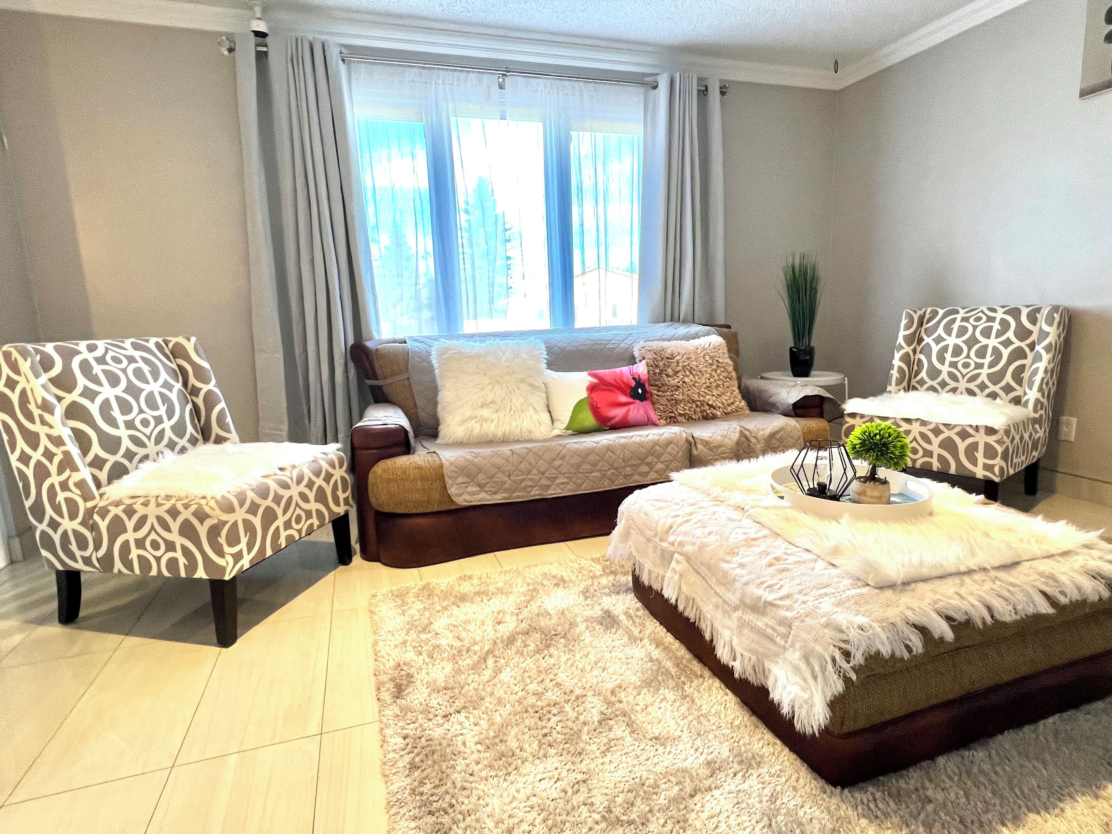 A living room with a sofa, two patterned armchairs, a coffee table, and a large window with curtains.