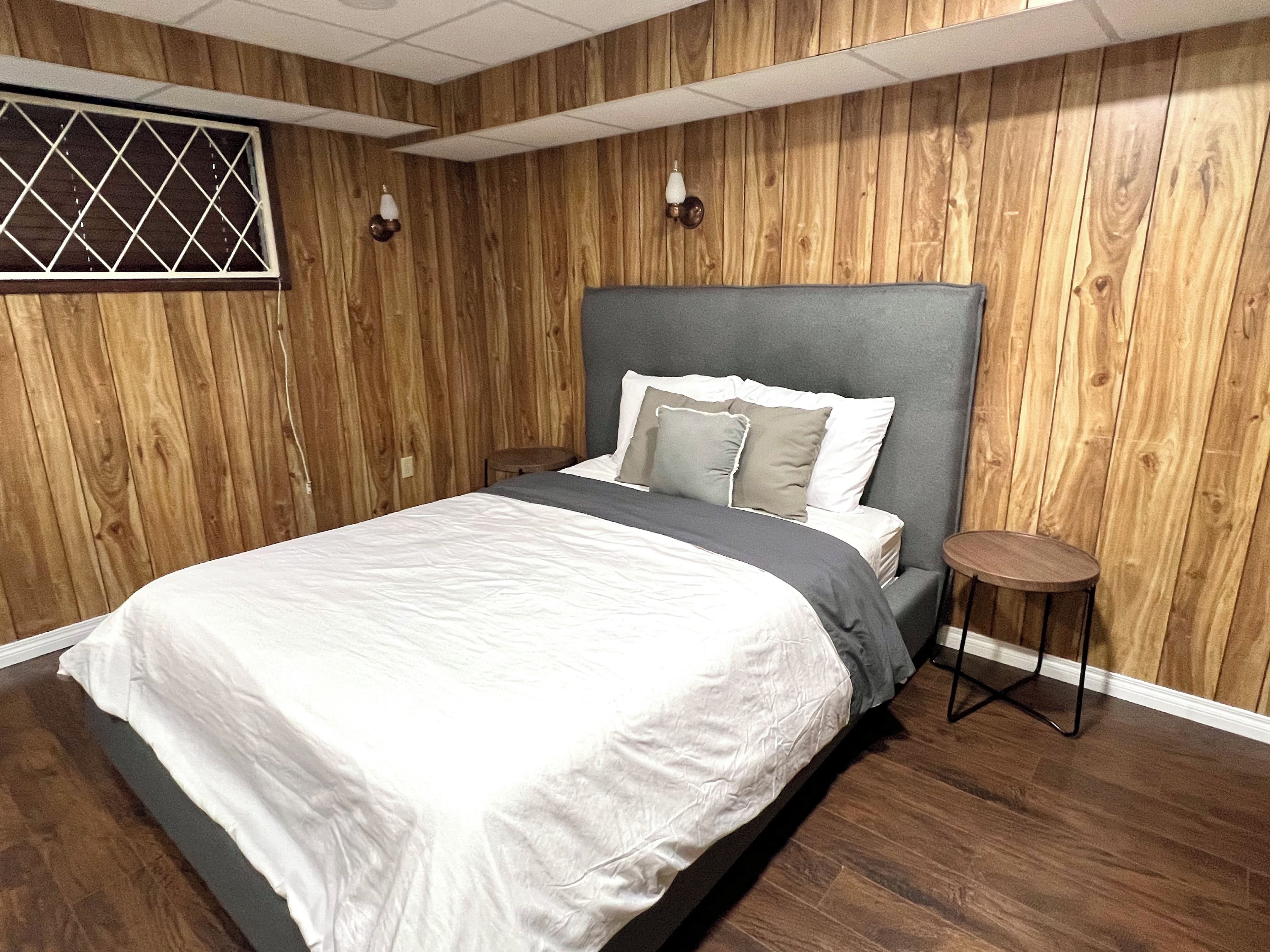 A bedroom with a wooden paneled wall, a bed with a grey headboard, and a small round table beside it.