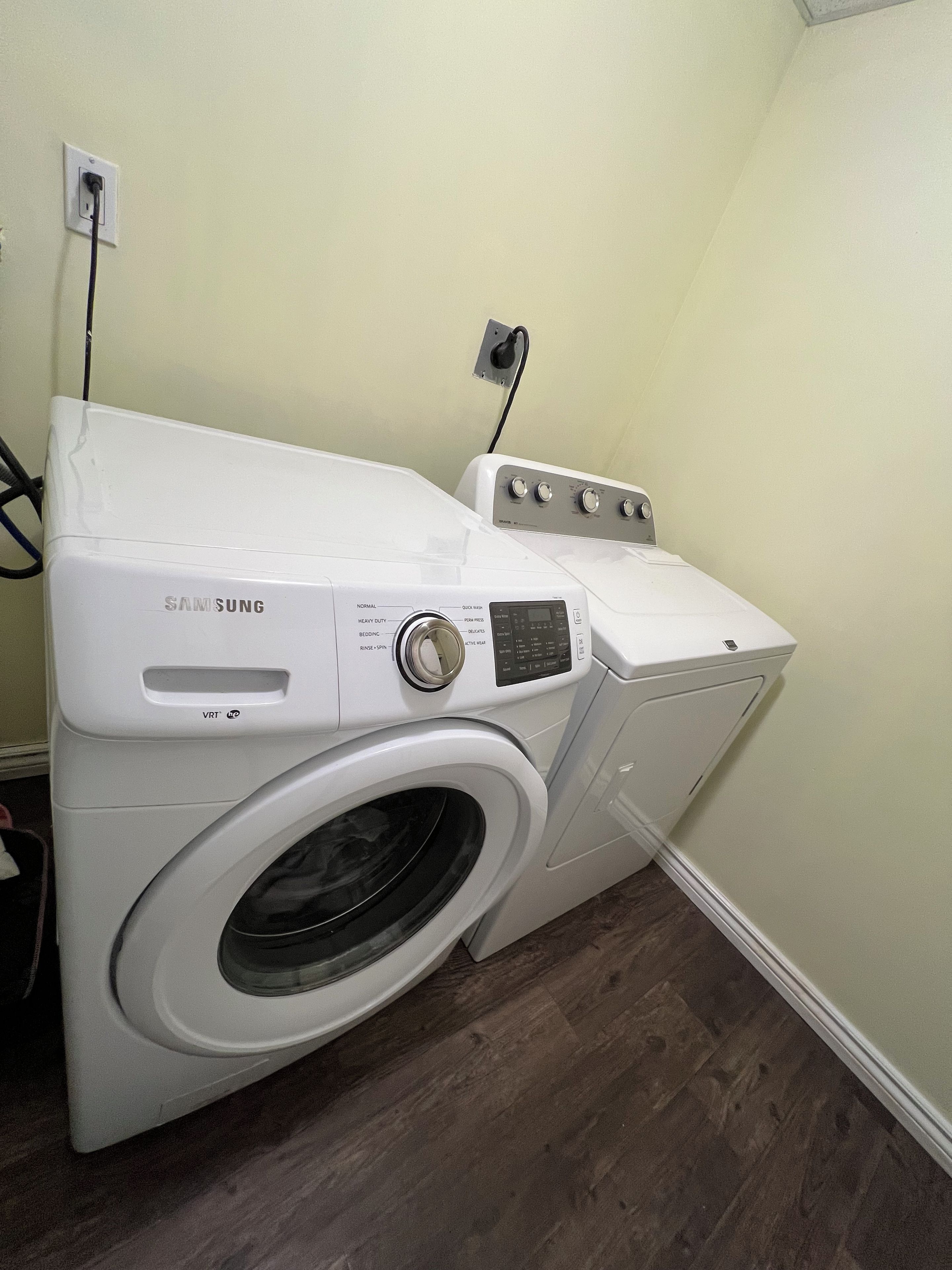 A front-loading Samsung washer and dryer unit in a laundry room with a wooden floor and a light-colored wall.