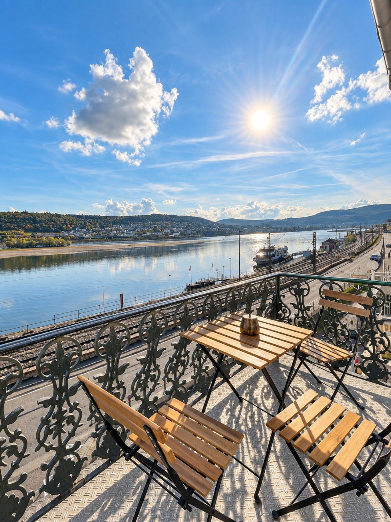 A riverside terrace with a table and benches, overlooking a calm river and distant hills.