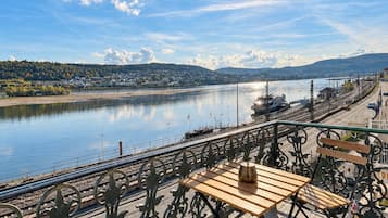 A riverside terrace with a table and benches, overlooking a calm river and distant hills.