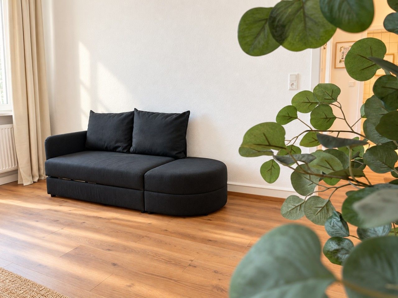 Un canapé noir avec deux coussins, dans une pièce dont le sol est recouvert de parquet et où se trouve une plante en pot.
