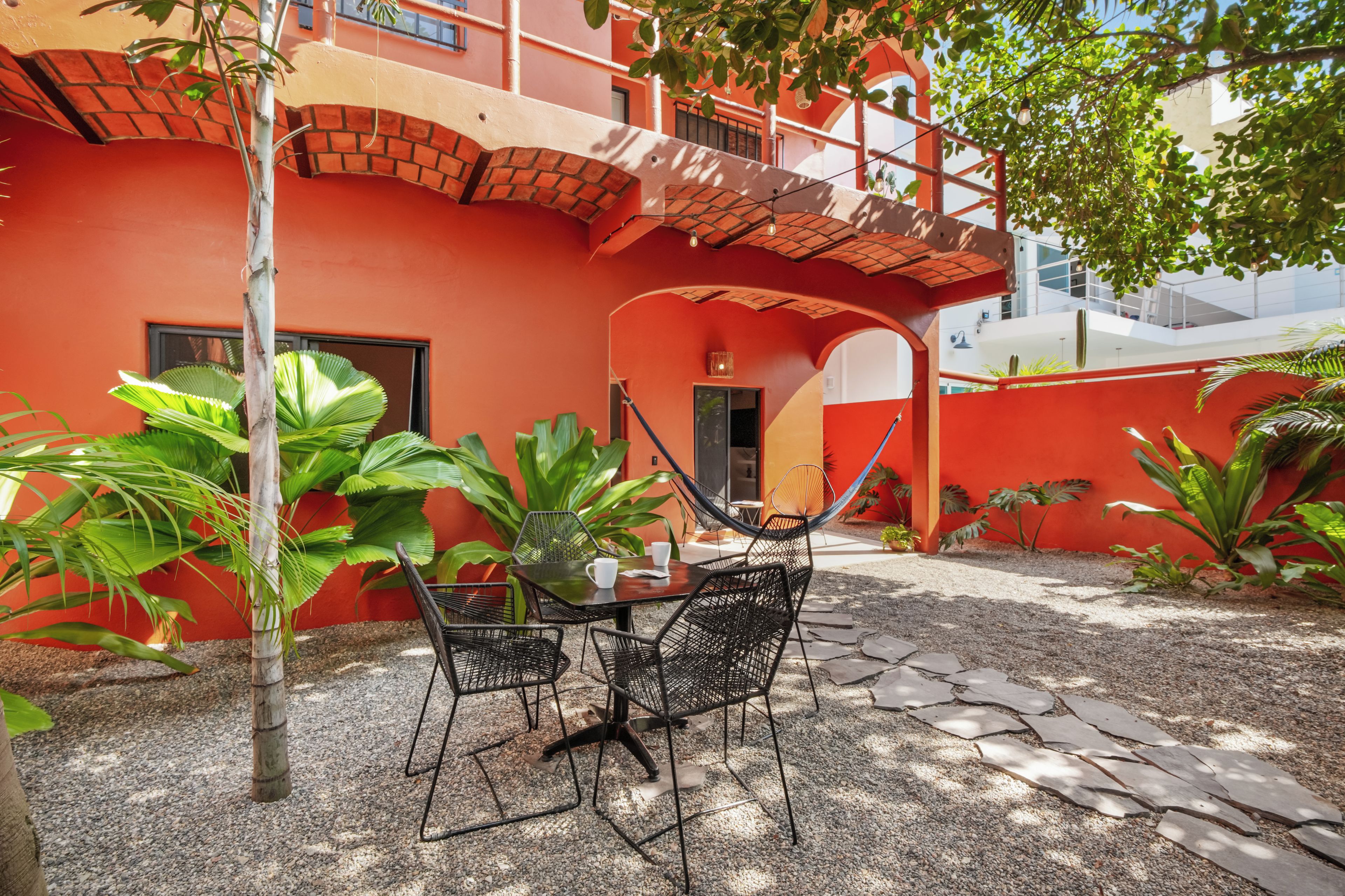A courtyard with a table and chairs, surrounded by plants and a red building with a brick archway.