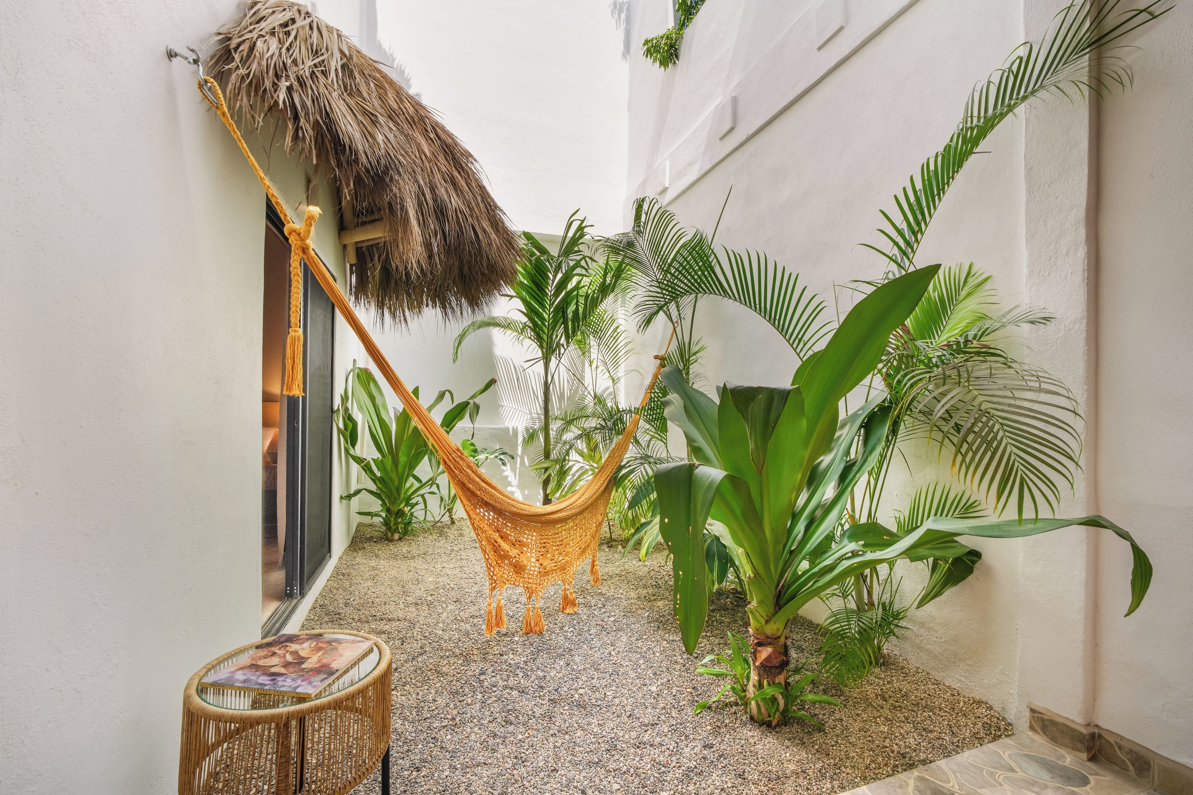 A narrow outdoor corridor with a hammock, potted plants, and a glass table.