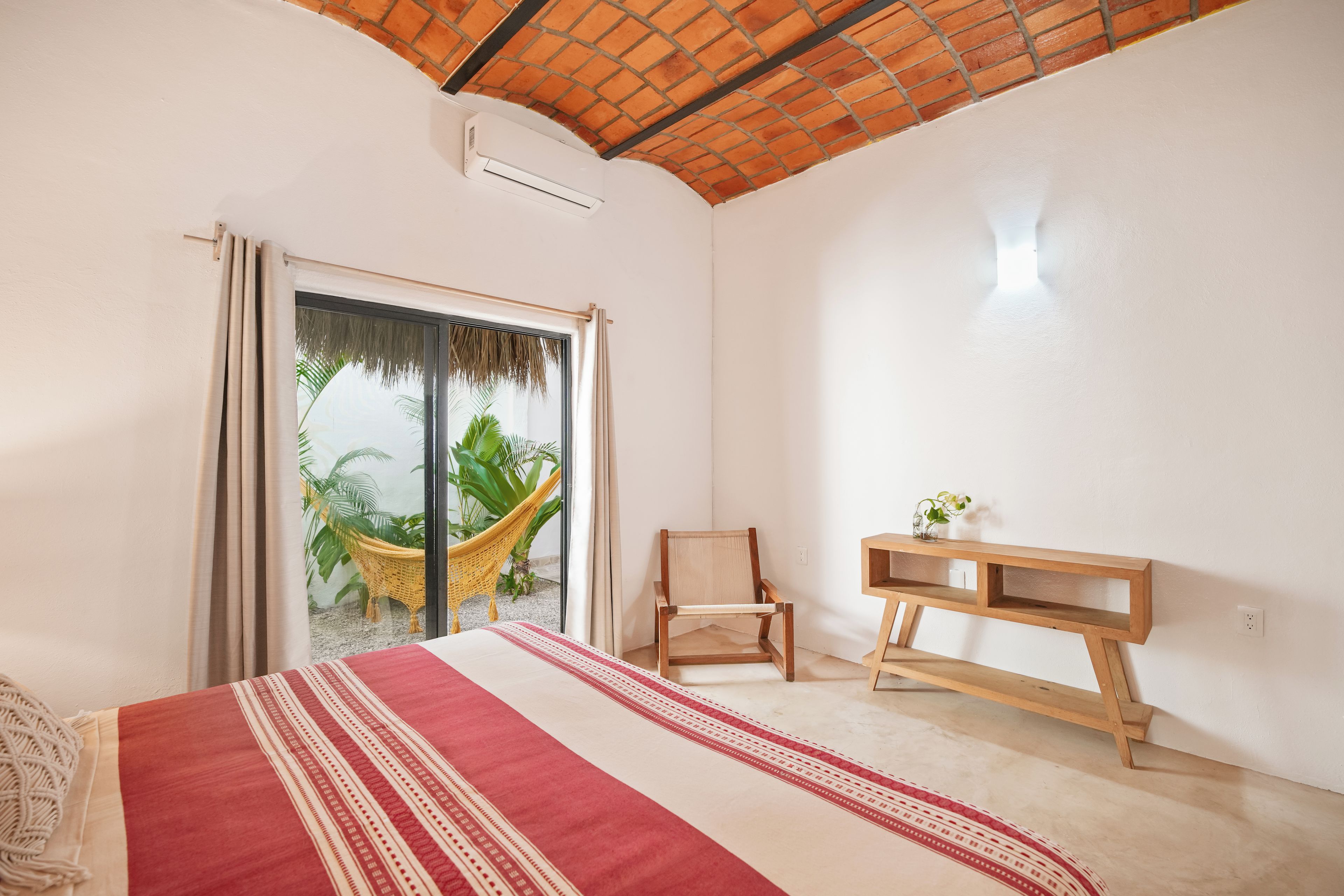 Un dormitorio con cama, una mesita de noche de madera, una silla, una hamaca y vistas a zonas verdes a través de una puerta corredera.