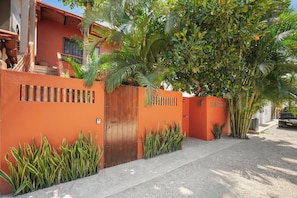 Una propietat residencial amb entrada tancada per reixa, un mur vermell i una gran varietat de plantes tropicals.