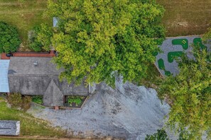 Une vue aérienne d’une propriété comprenant un grand arbre, un bâtiment doté d’un toit en pente et une zone recouverte de gravier avec des zones d’herbe.