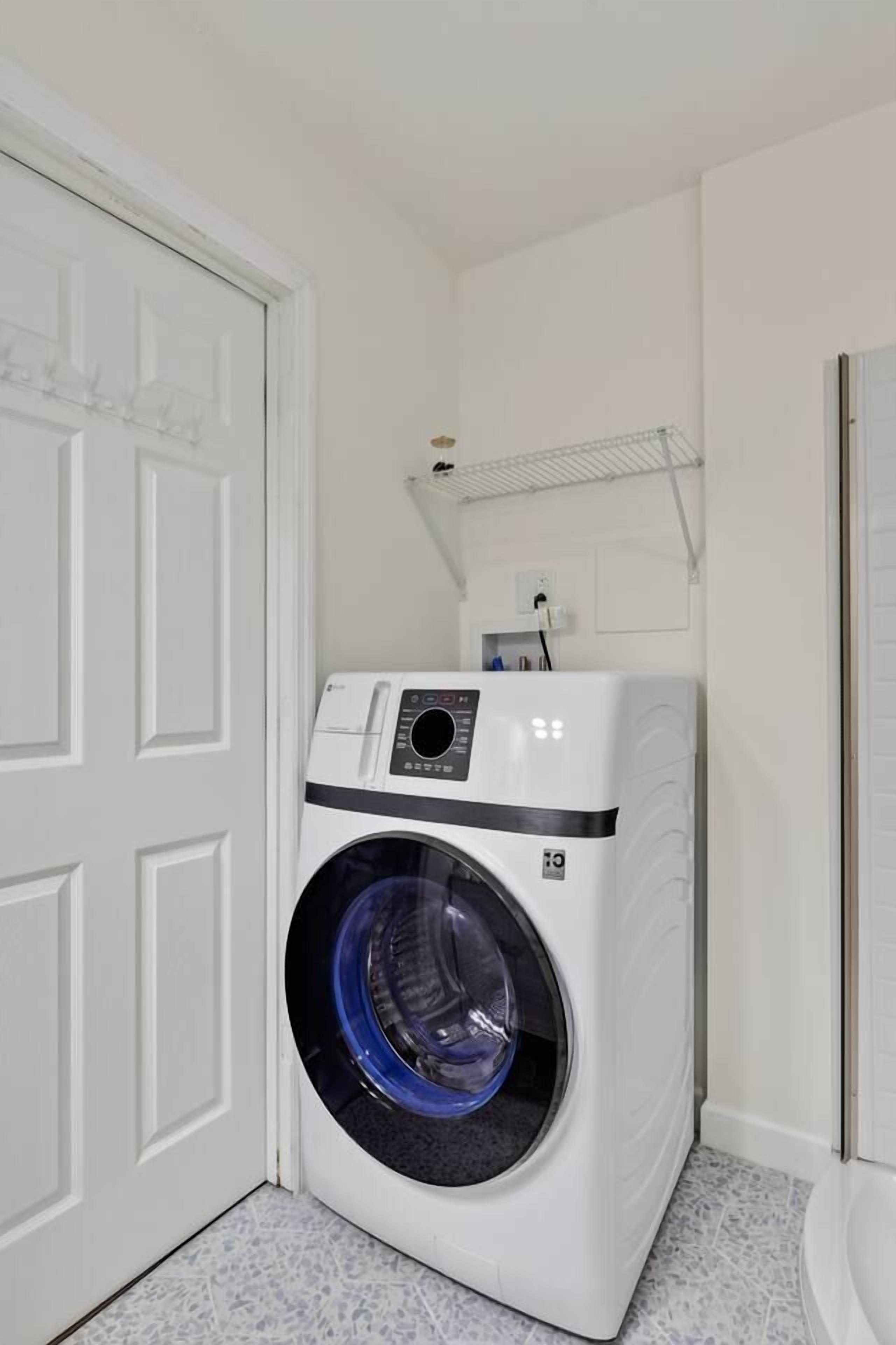 A compact, white front-loading washer and dryer unit in a small laundry room with a white door and tiled floor.
