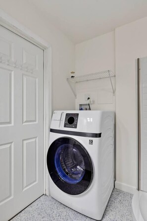 A compact, white front-loading washer and dryer unit in a small laundry room with a white door and tiled floor. - 2-Cabin Riverfront Compound: Pickleball+Mini Golf (Shenandoah)