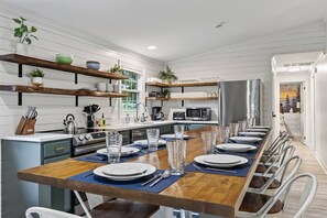 A modern kitchen with a long dining table set for six, equipped with a microwave, stove, and various utensils. - 2-Cabin Riverfront Compound: Pickleball+Mini Golf (Shenandoah)