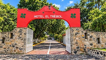 Un arc rouge surmonté de l’inscription « HÔTEL EL TRÉBOL », qui mène à un chemin pavé bordé de murs de pierre et d’arbres.