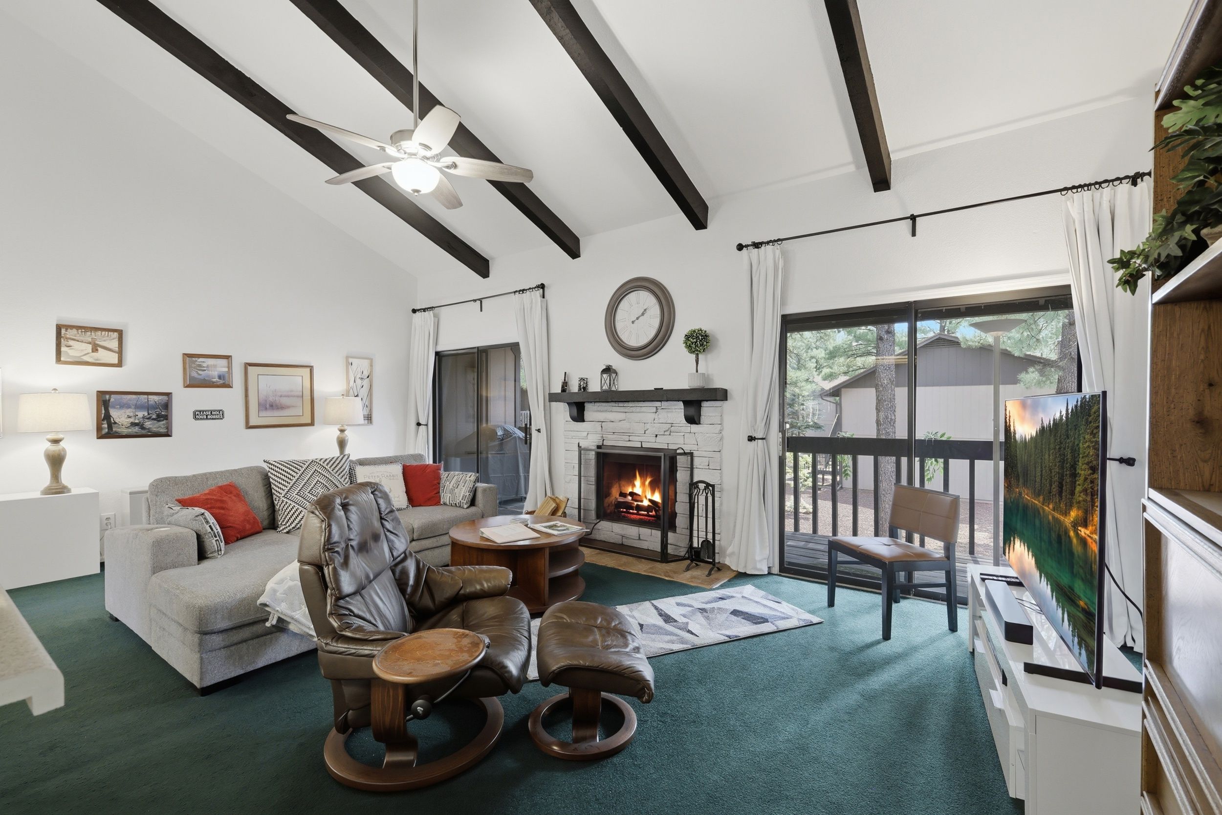 A living room with a fireplace, a ceiling fan, and a view of a forest through a glass door.