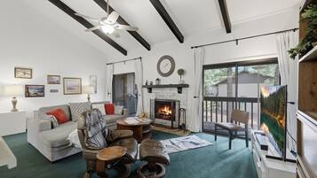 A living room with a fireplace, a ceiling fan, and a view of a forest through a glass door.