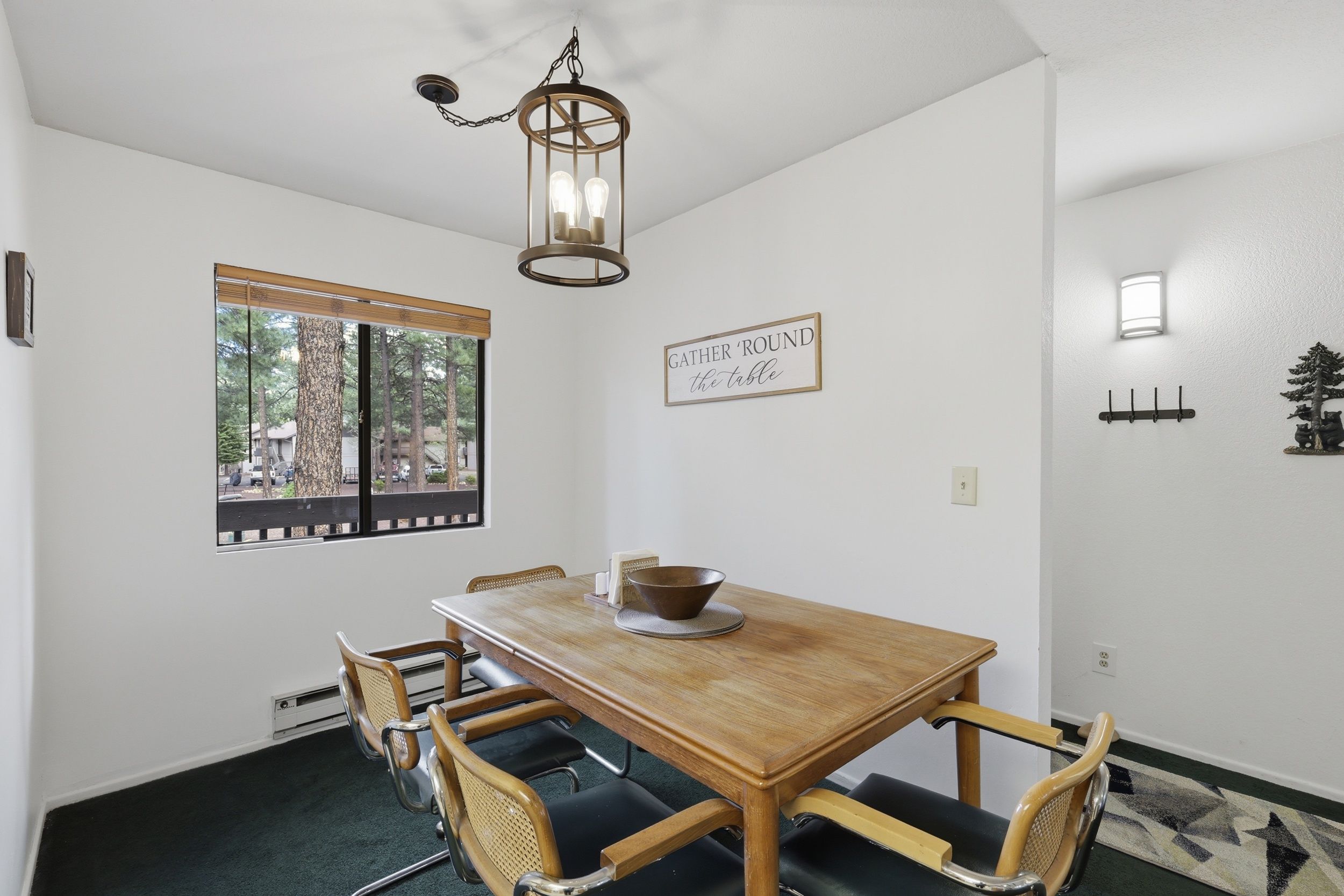 A dining room with a wooden table, four chairs, a hanging light fixture, a window with a view, and a wall-mounted sign.