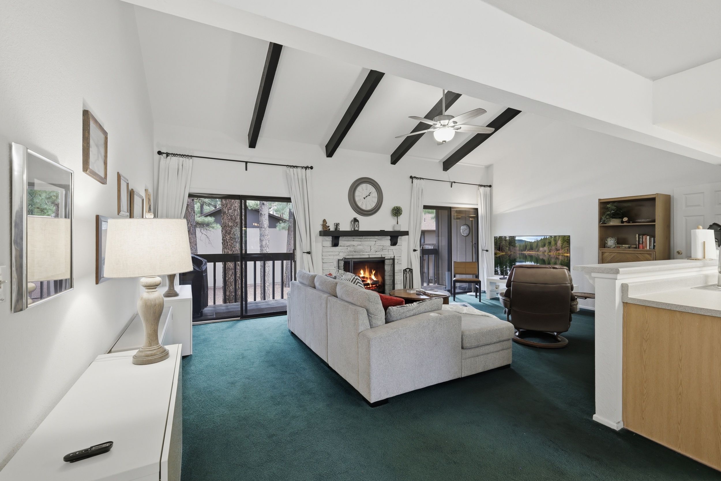 A spacious living room with a fireplace, a sofa, a coffee table, and a television. There is a balcony with a view of trees and a pool.