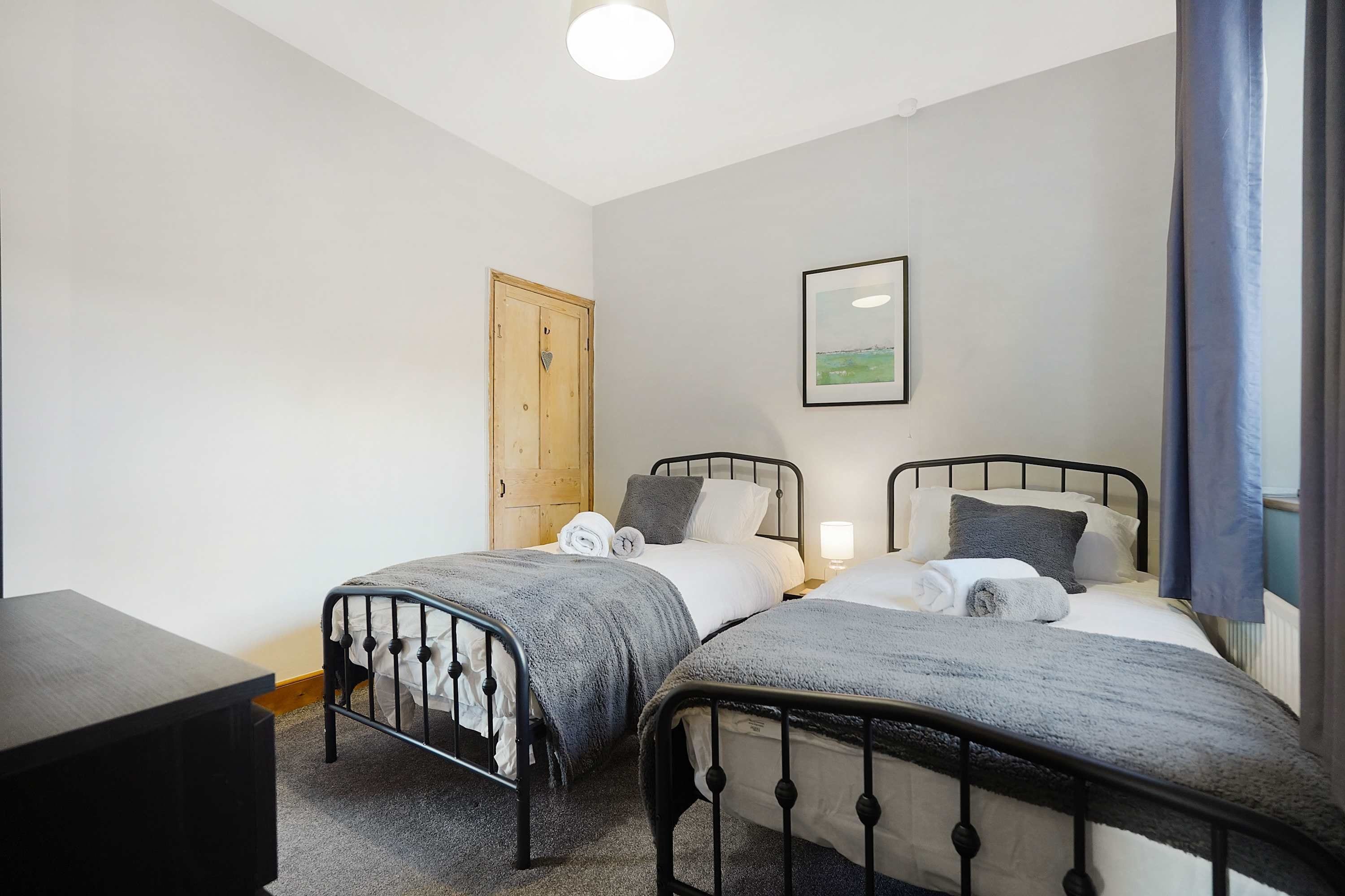 Habitación doble con dos camas, una puerta de madera, un cuadro enmarcado en la pared y una mesita de noche con una lámpara.