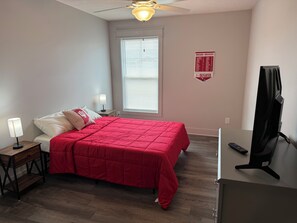 Une chambre à coucher comprenant un lit, une table de chevet, une lampe, un ventilateur de plafond, une fenêtre avec des stores, un téléviseur et un panneau fixé au mur.