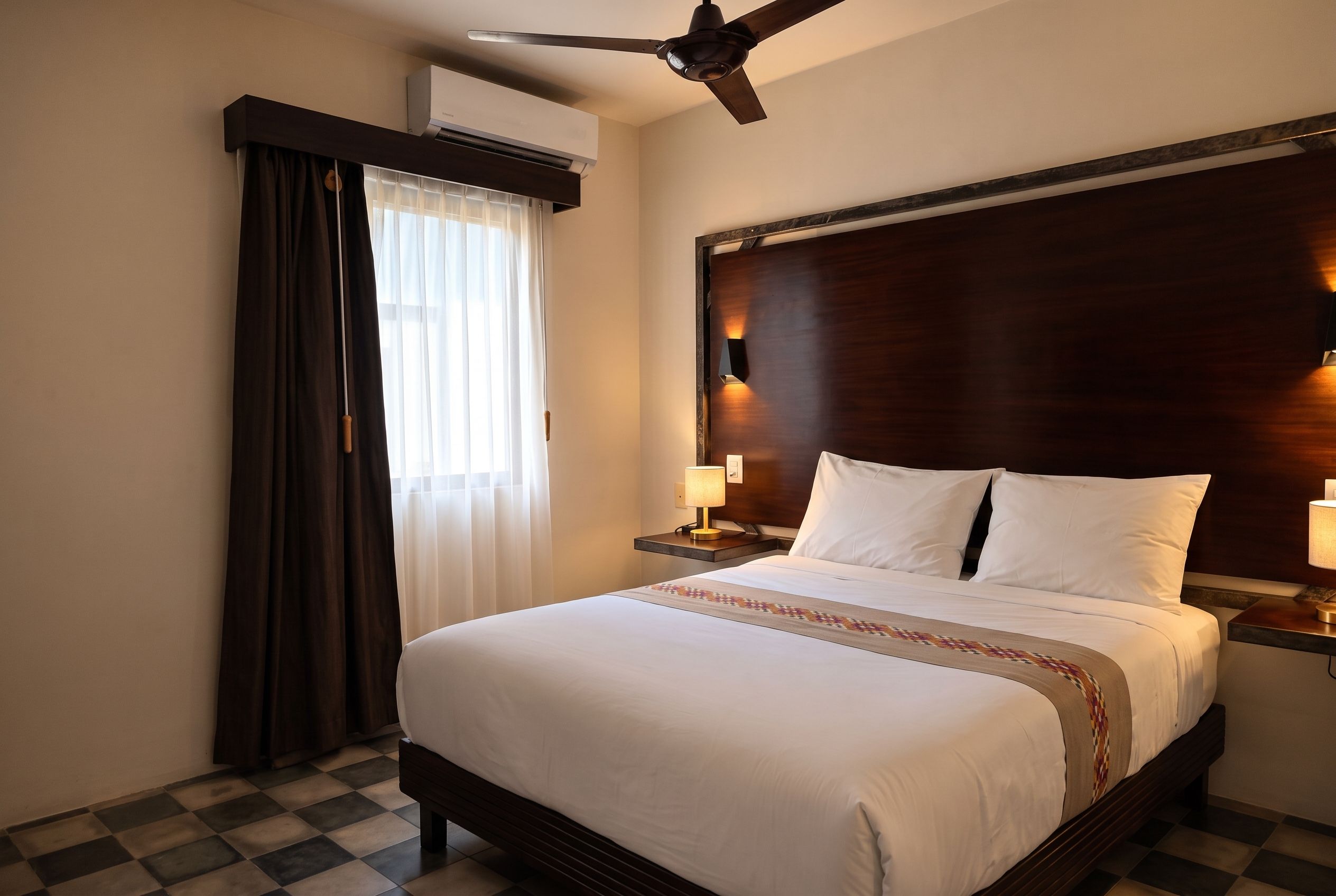 Habitación de hotel con cama, ventilador de techo, unidad de aire acondicionado, mesita de noche con lámpara y ventana con cortinas.