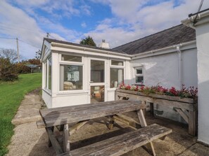 A white cottage with a conservatory, a wooden bench, and a flower bed. - BRYN EITHIN, family friendly, country holiday cottage in Carmel (Caernarfon)