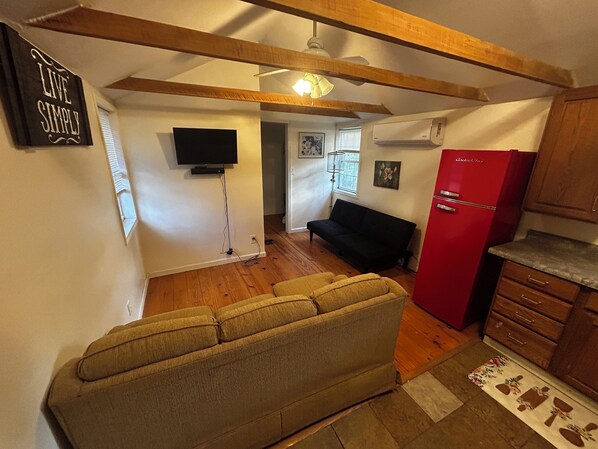 A compact living space with a sofa, a flat-screen TV mounted on the wall, a red refrigerator, and wooden beams on the ceiling.