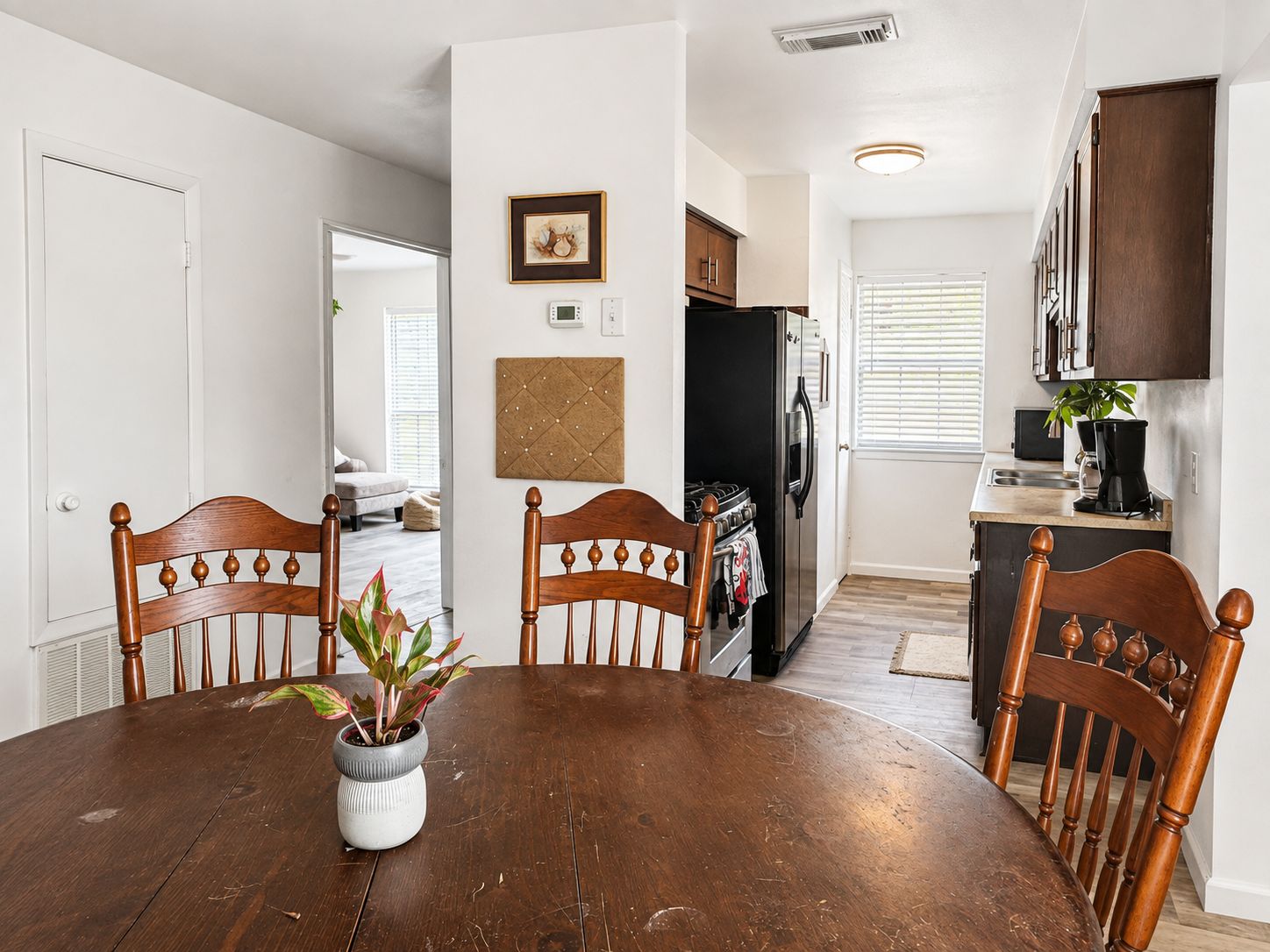 A dining area with a wooden table and chairs, a potted plant, and a kitchen in the background.