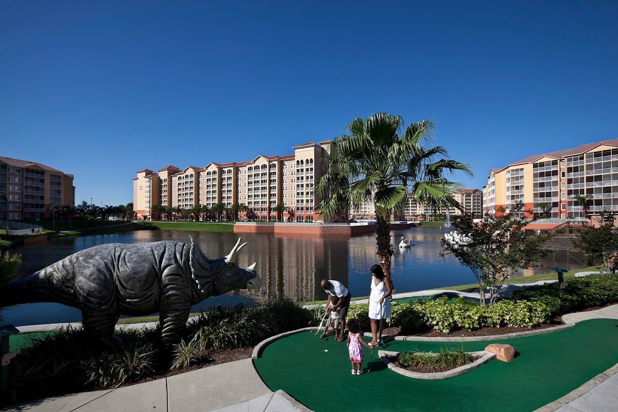 Een golfbaan met een putting green, een groot dinosaurusbeeld en een hotel op de achtergrond.
