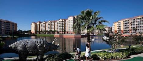 Een golfbaan met een putting green, een groot dinosaurusbeeld en een hotel op de achtergrond.