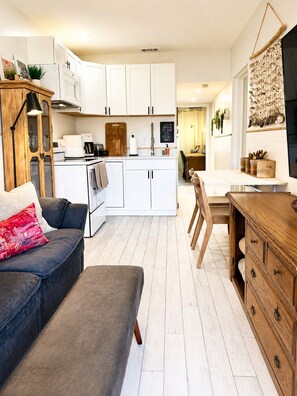 Un espacio habitable compacto con kitchenette, comedor y zona de estar con sofá y reposapiés.