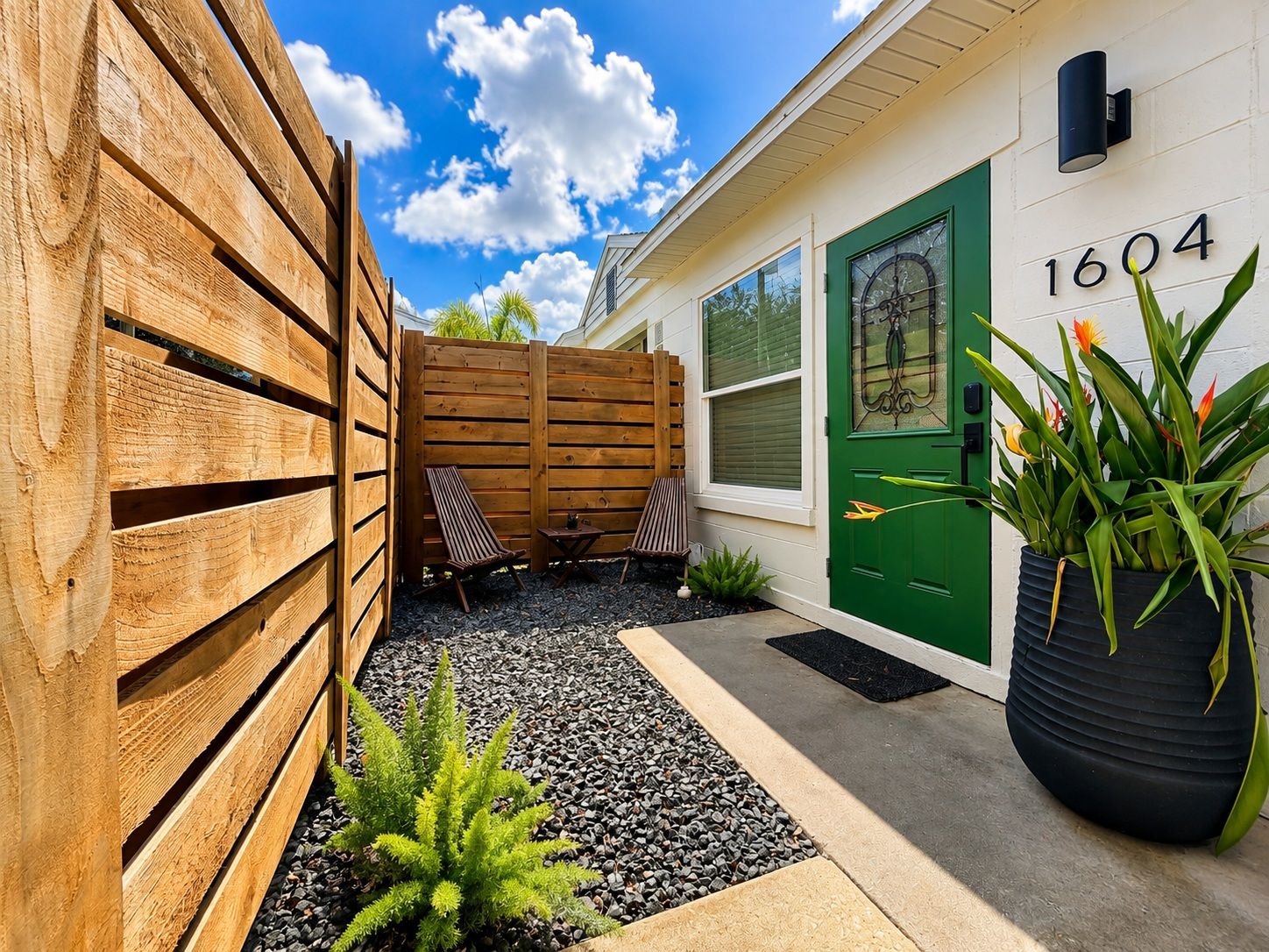 A residential property with a green door marked '1604,' a wooden fence, outdoor seating, and a potted plant.