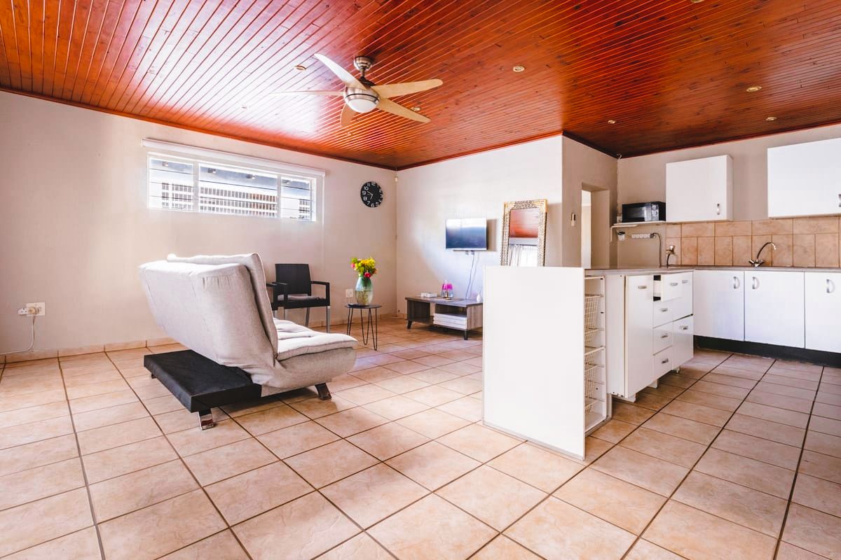 Een ruime kamer met een plafondventilator, een bed, een klein tafeltje met een vaas en een kitchenette.