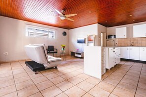 Una habitación amplia con ventilador de techo, una cama, una mesita con un florero y una zona de kitchenette.