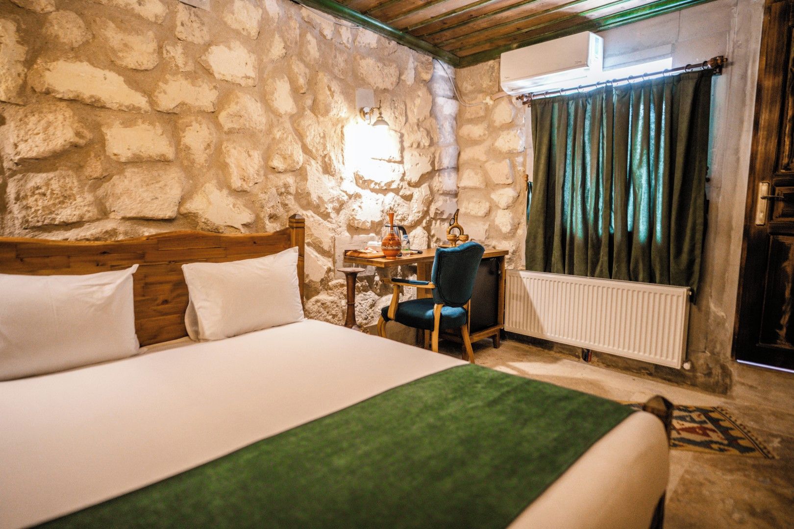A room with a stone wall, a bed with white bedding, a wooden headboard, a desk with a chair, a radiator, and a window with curtains.