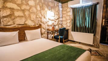 A room with a stone wall, a bed with white bedding, a wooden headboard, a desk with a chair, a radiator, and a window with curtains.