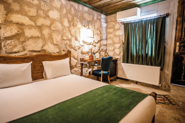 A room with a stone wall, a bed with white bedding, a wooden headboard, a desk with a chair, a radiator, and a window with curtains.