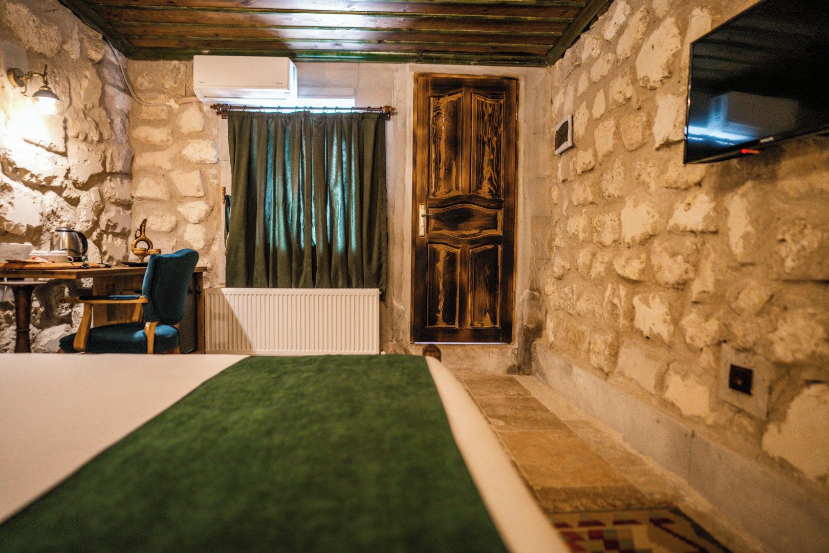 A room with a bed, a chair, a table with a kettle, a radiator, a wooden door, and a television mounted on the wall.