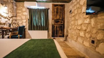 A room with a bed, a chair, a table with a kettle, a radiator, a wooden door, and a television mounted on the wall.