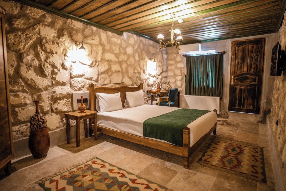 A bedroom with a stone wall, a wooden bed, a nightstand, a vase, a chandelier, and a patterned rug.