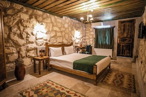 A bedroom with a stone wall, a wooden bed, a nightstand, a vase, a chandelier, and a patterned rug. - Vista Rosa Cappadocia (Çavuşin)