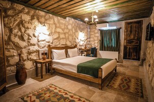 Un dormitorio con pared de piedra, una cama de madera, una mesita de noche, un florero, una araña y una alfombra estampada.