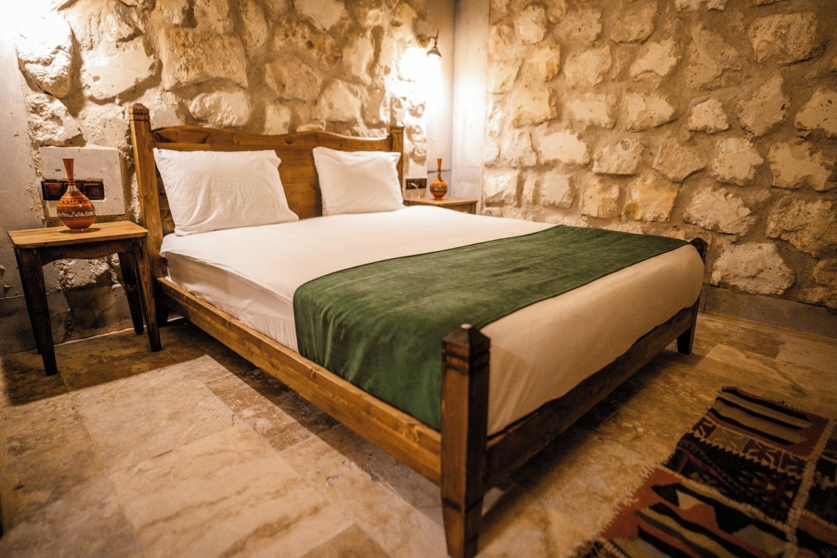 A room with a stone wall, a wooden bed with white bedding and a green throw, a nightstand with a vase, and a rug.